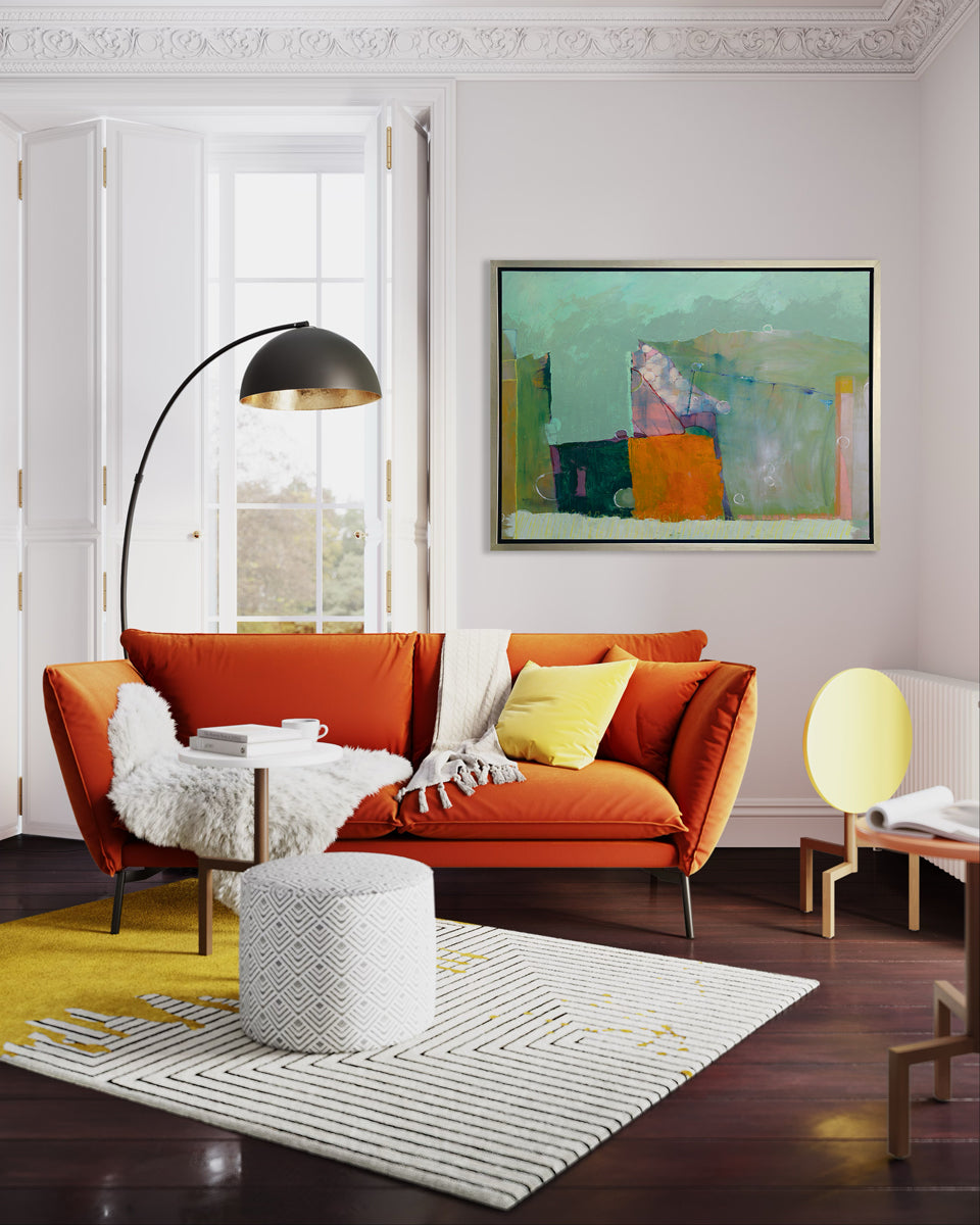A teal, orange and green abstract print in a silver floater frame hangs on a white wall above a dark orange sofa with pillows and white throw blankets, a large floor lamp and a patterned ottoman on top of a patterned area rug.