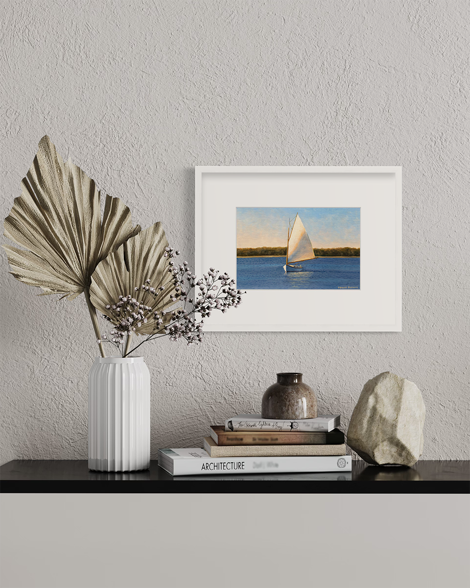 A blue and beige contemporary seascape print with a sailboat in a white frame with a mat hangs on a white wall above a black surface with a stack of books, a rock and a white vase with decorative stems.