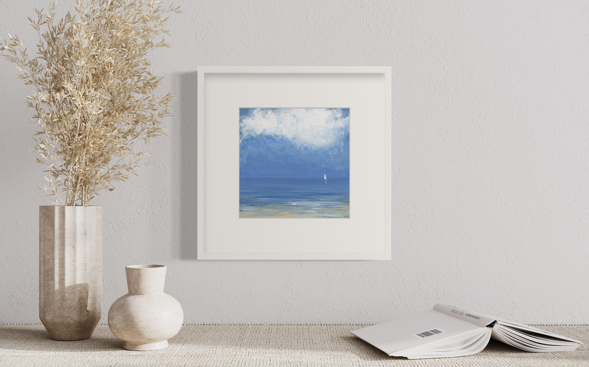 A blue, beige and white abstract seascape print with a small sailboat in a white frame with a mat hangs on a white wall above an open book and two vases.