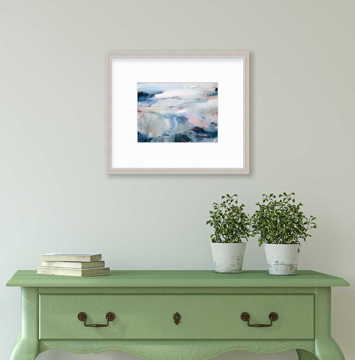A blue abstract print in a silver frame with a mat hangs on a white wall above a green console table with a stack of books and two potted plants.