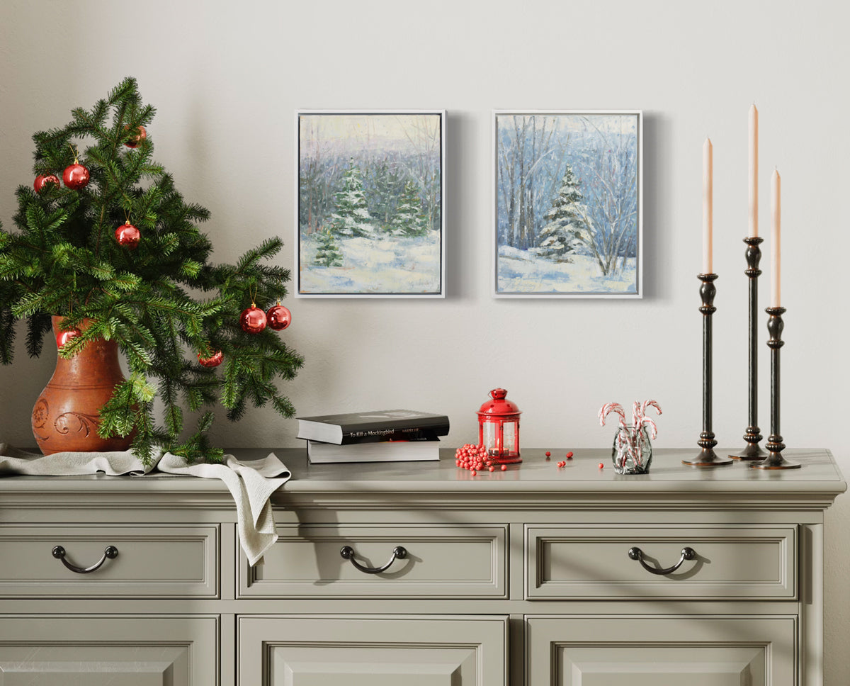 Two blue, white and green snow fall landscape paintings with snow covered pine trees hang in white floater frames on a white wall side by side above a beige credenza with candles, books and some festive decorations.