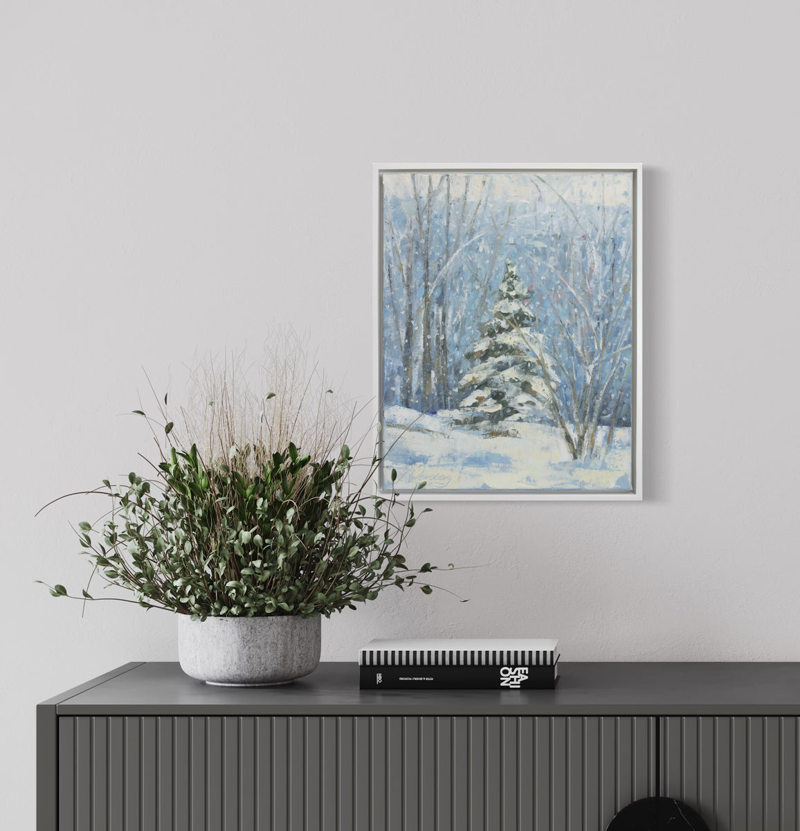 A blue, white and green snow fall landscape painting with one snow covered pine tree in the center by Molly Doe Wensberg hangs in a white floater frame on a white wall above a grey credenza with a potted plant and a stack of books.