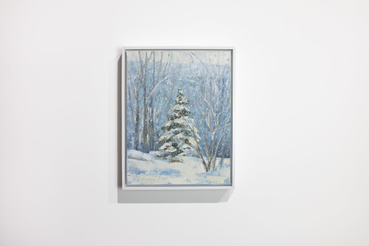 A blue, white and green snow fall landscape painting with one snow covered pine tree in the center by Molly Doe Wensberg hangs in a white floater frame on a white wall.