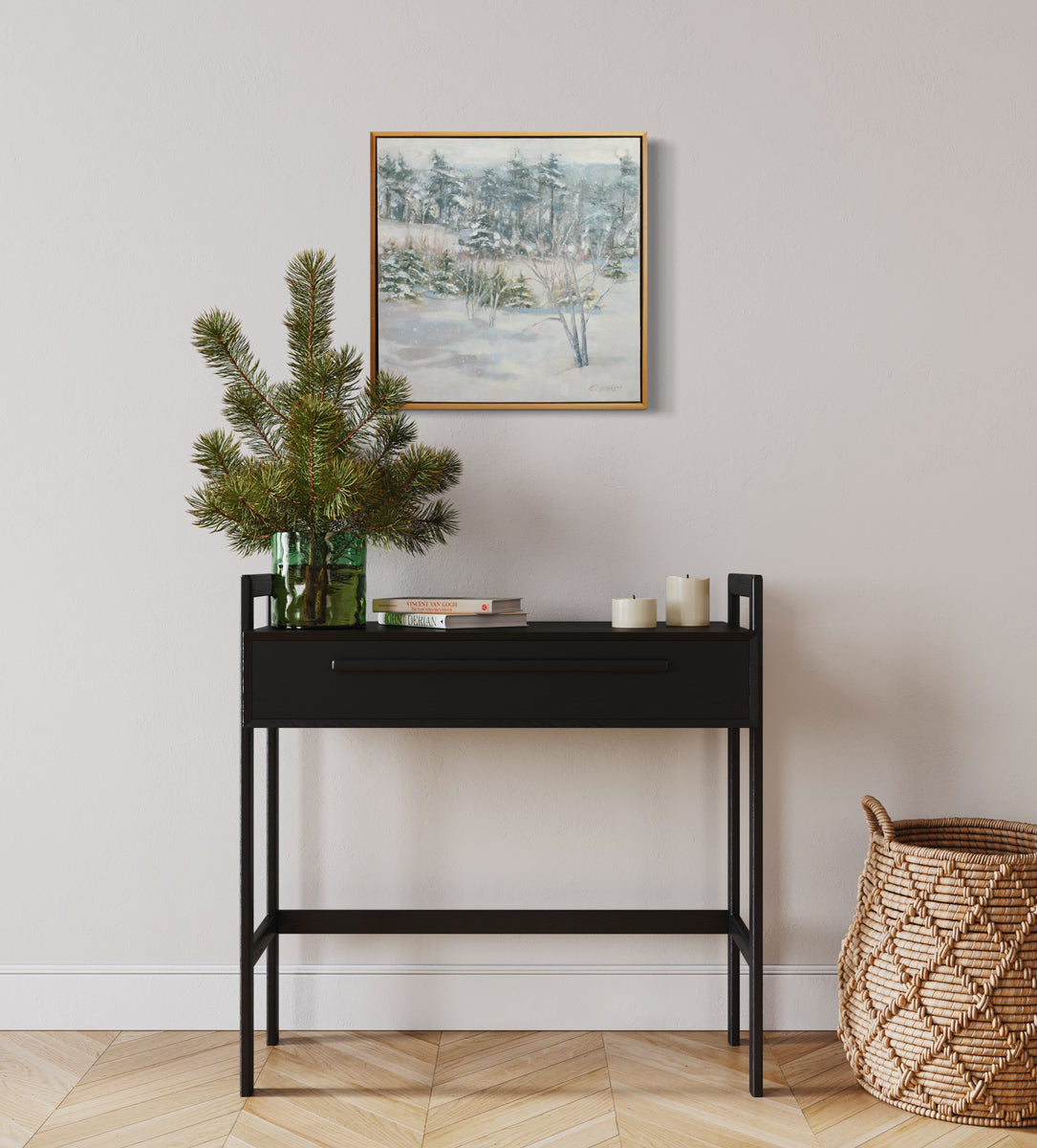 A white, grey and green snow landscape painting by Molly Doe Wensberg hangs in a light wood floater frame on a white wall above a black table with candles, books and a vase with pine branches.