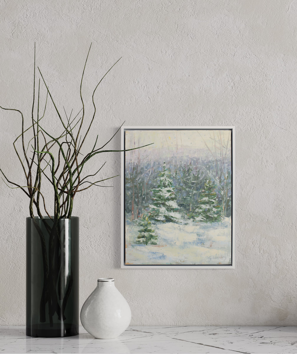 A blue, white and green snow fall landscape painting with snow covered pine trees in the center by Molly Doe Wensberg hangs in a white floater frame on a white wall above a white marble surface with a white mini vase and a dark cylindrical vase with stems.