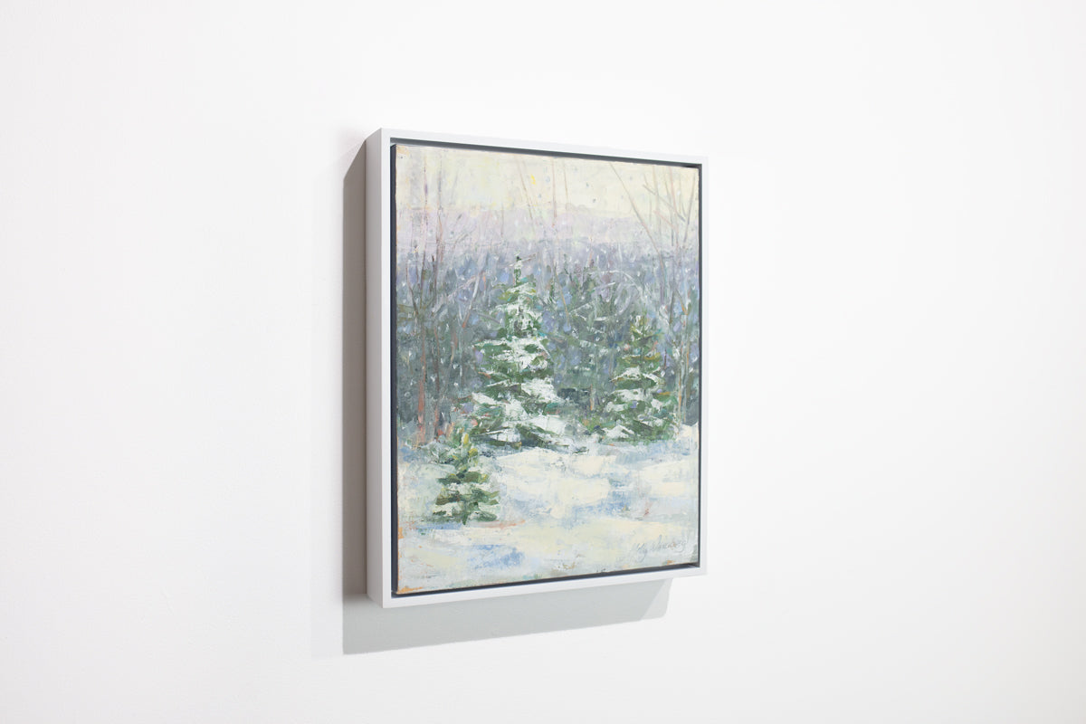 A side view of a blue, white and green snow fall landscape painting with snow covered pine trees in the center by Molly Doe Wensberg hangs in a white floater frame on a white wall.