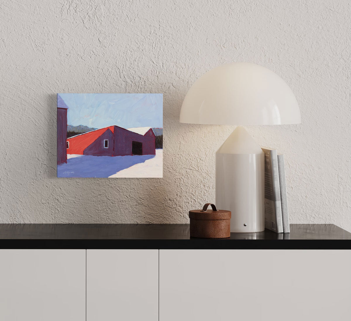 A small landscape painting of a red barn in snow hangs on a textured wall above a black and white console table and a modern white table lamp, books, and a small round box. 