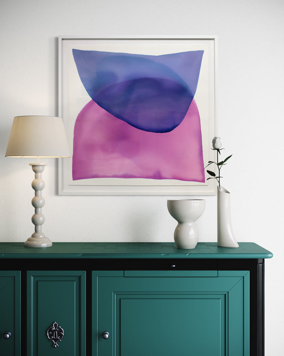 A blue and magenta minimalist abstract watercolor painting framed in a white frame hangs on a white wall above a teal console table with white ceramic vases and a white table lamp. 