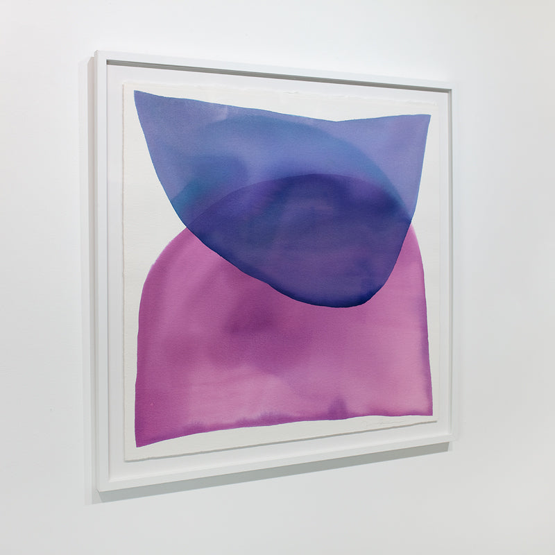 A blue and magenta minimalist abstract watercolor painting framed in a white frame hangs on a white wall.