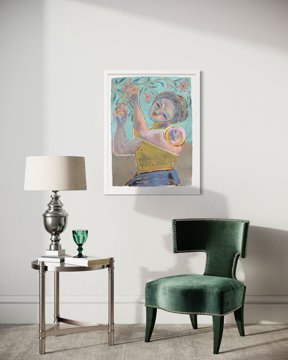 A framed abstracted blue, turquoise and yellow painting of a woman holding flowers from a branch hangs on a white wall above a modern green velvet armchair and a silver end table with books and a silver lamp and green glass. 