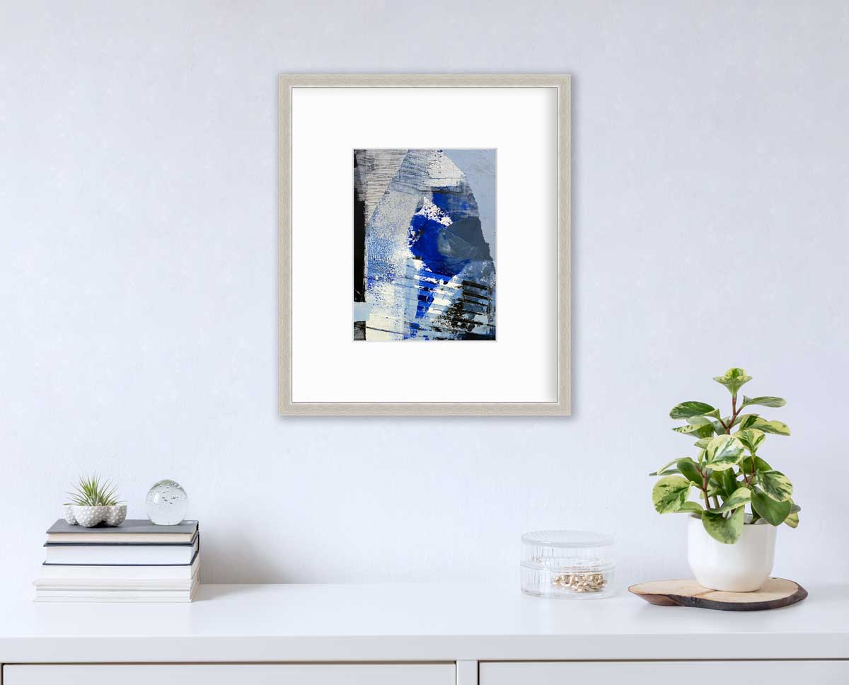 A blue, white and black abstract print in a silver frame with a mat hangs on a white wall above a white surface with a stack of books, a round clear container with gold accessories and a potted plant.