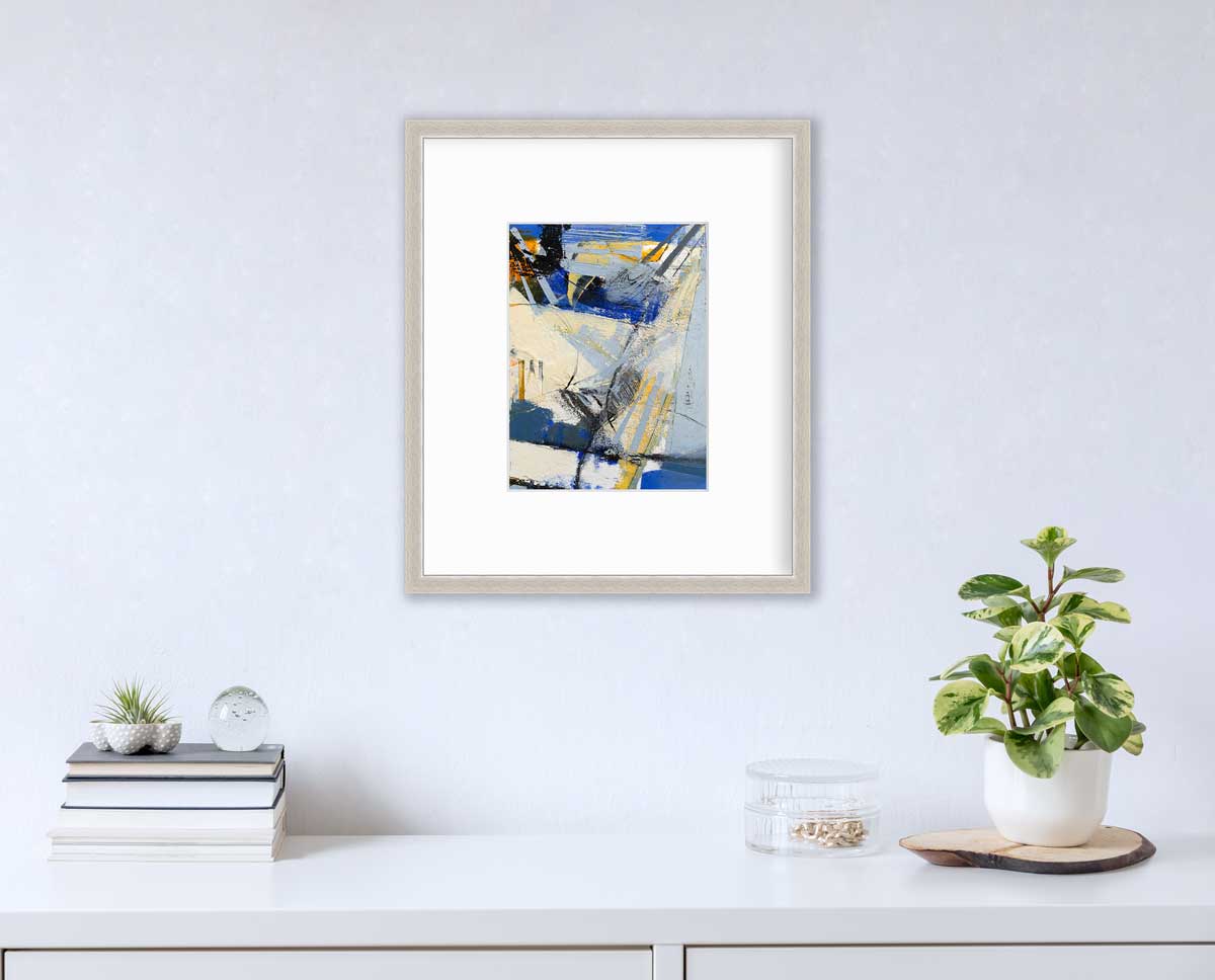 A blue, white, yellow and black abstract print in a silver frame with a mat hangs on a white wall above a white surface with a stack of books, a round clear container with gold accessories and a potted plant.