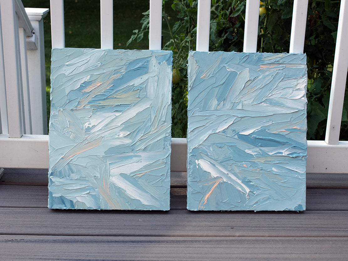 A pair of thickly painted paintings in teal, sea foam green, celadon, white and hints of orange and yellow by Teodora Guererra leaning on a deck outside. The two painting are like wall sculptures.
