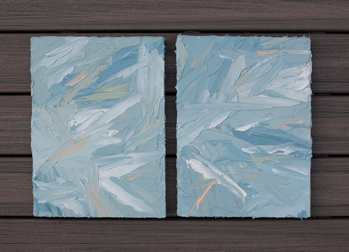 A pair of thickly painted paintings in teal, sea foam green, celadon, white and hints of orange and yellow by Teodora Guererra laying flat on a deck outside. The two painting are like wall sculptures.