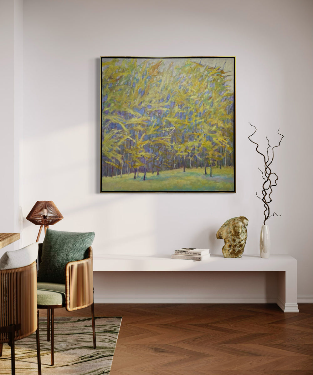 An impressionistic painting similar to artist Wolf Kahn of a forest with bright yellow leaves and a lavender grey background hangs in a wam silver floater frame over a white bench with decorative adornments.