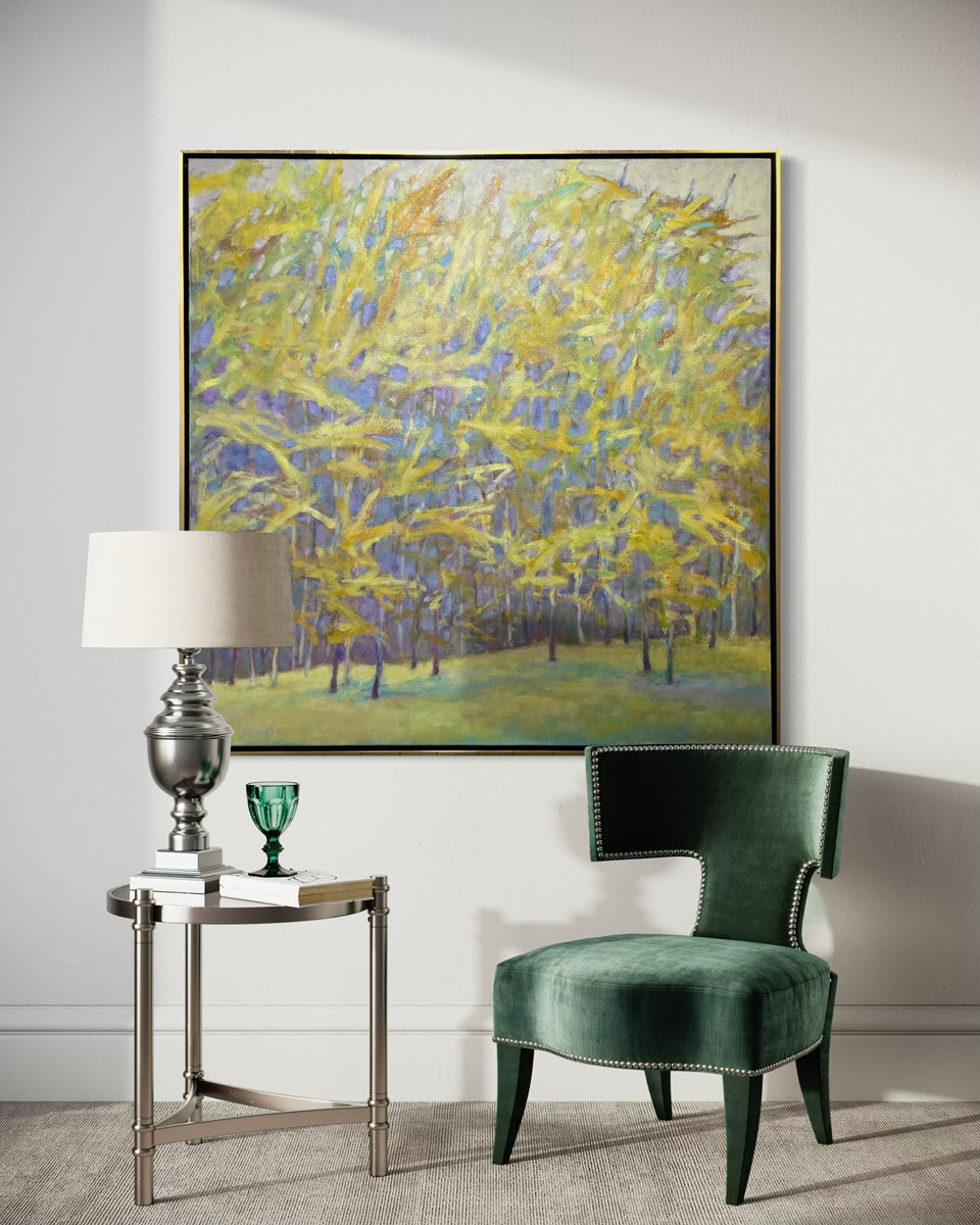 An impressionistic painting similar to artist Wolf Kahn of a forest with bright yellow leaves and a lavender grey background hangs in a wam silver floater frame on a white wall with a velvet green chair in front.