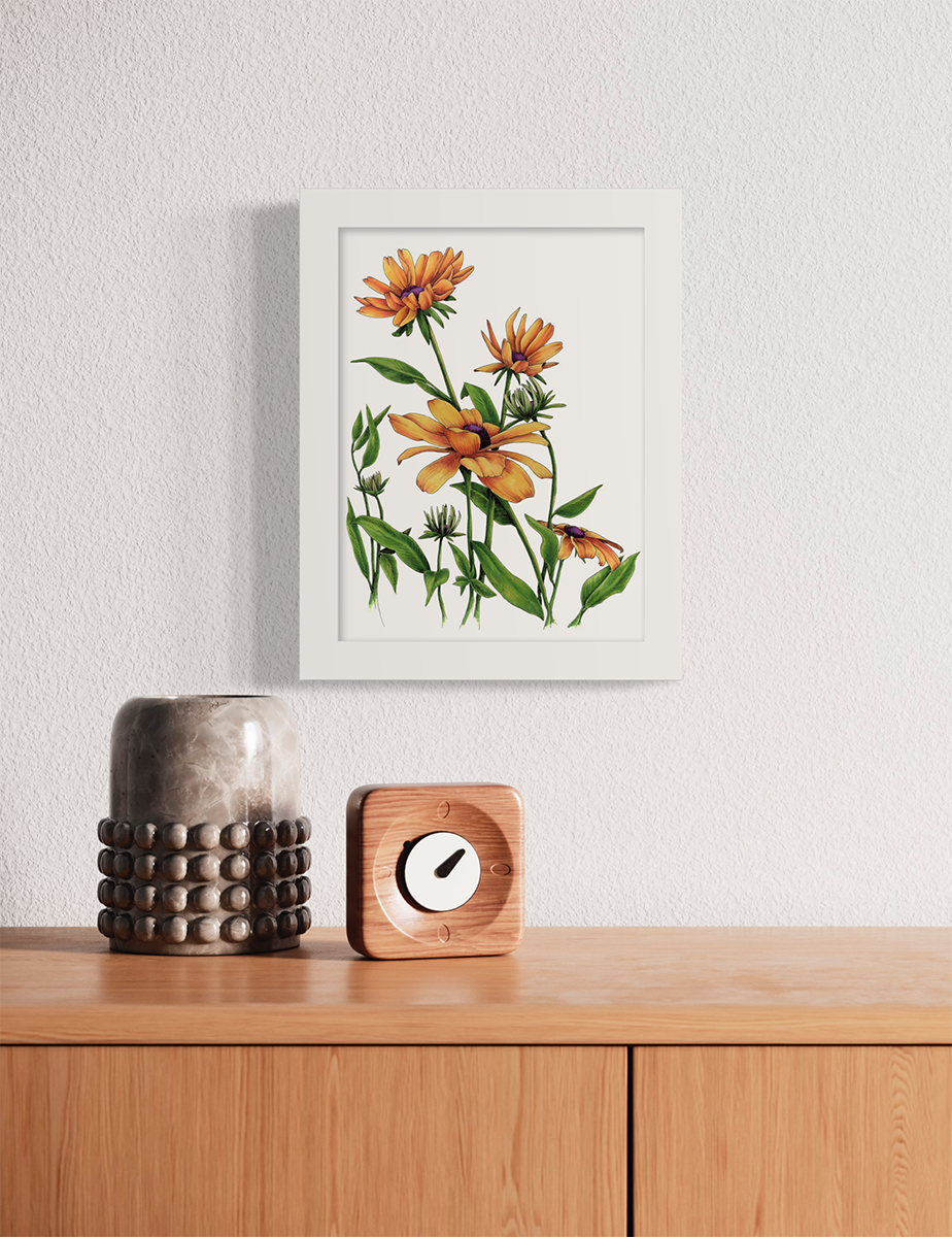 Yellow and orange flowers with a purple center and green stems and leaves print in a white frame hangs on a white wall above a wood surface with a small wood analog clock and a grey candleholder.
