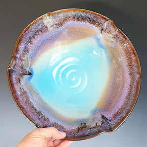 A hand holds a multicolored ceramic serving bowl in front of a grey gradient backdrop. 