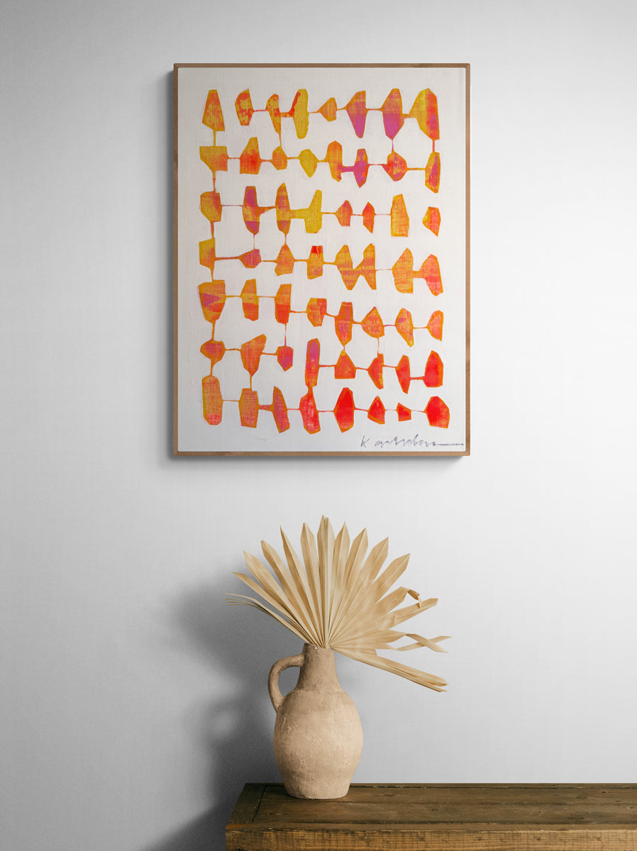 Framed abstract art with colorful patterns on a wall above a wooden shelf with a vase and dried plant.