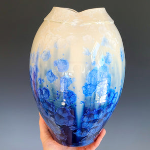 A hand holds a blue and cream-colored glazed ceramic vase with a wide neck in front of a grey backdrop. 