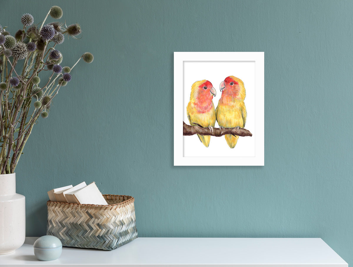 An illustration of two yellow birds on a branch framed in a white frame hangs on a turquoise wall above a white table with woven and ceramic table decor. 