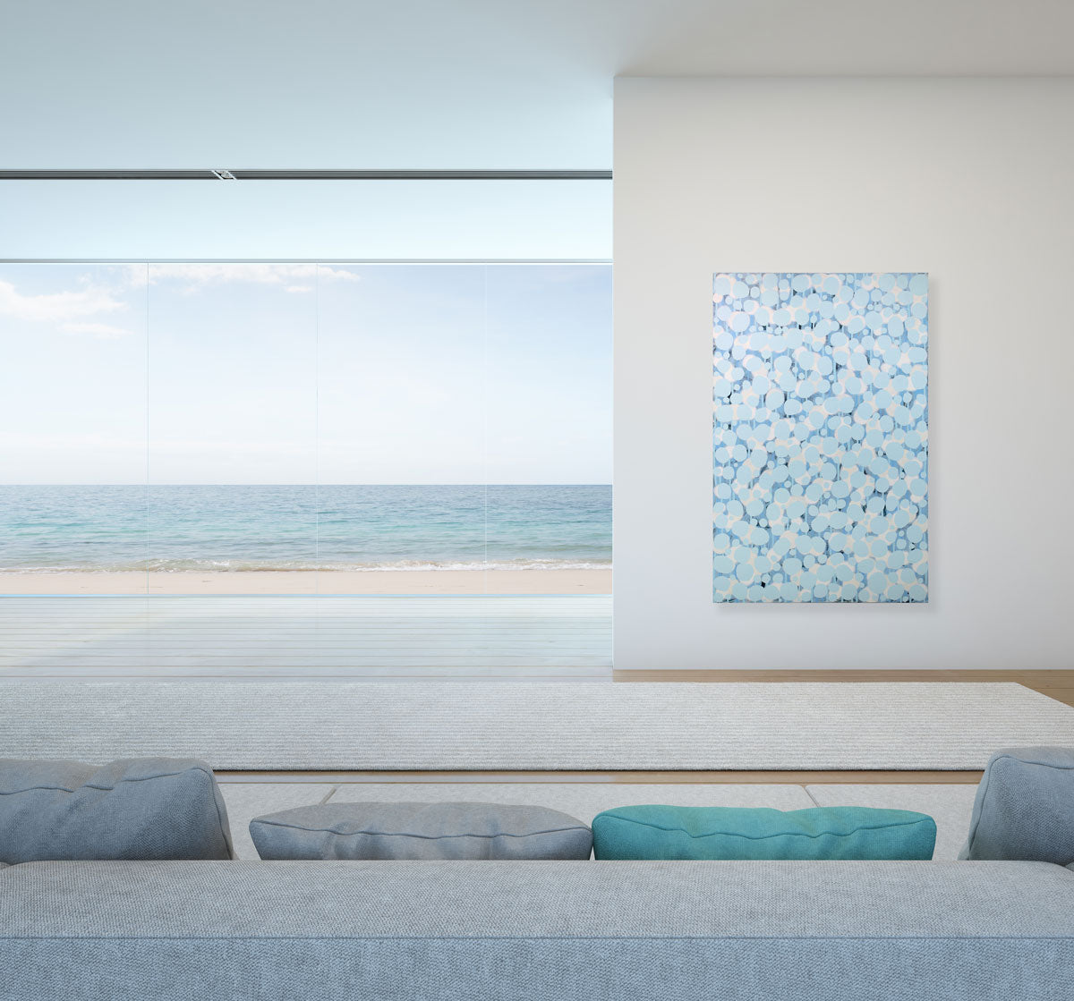 An abstract blue and white painting hangs on a white wall next to a large floor-to-ceiling window overlooking the ocean beach. 