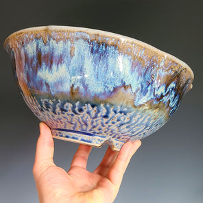 A man's hand holds a blue and brown ceramic bowl in front of a grey ombre backdrop. 