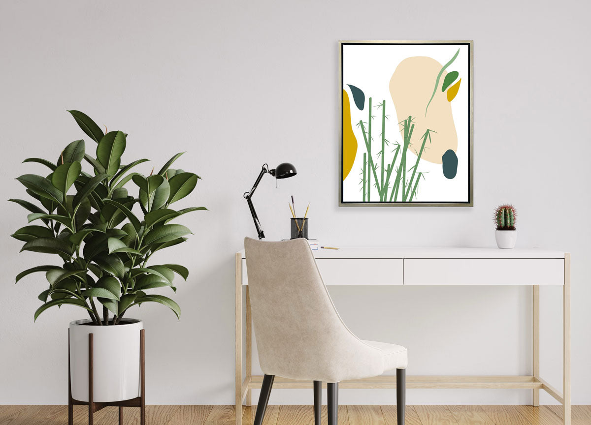 An abstract yellow, beige, blue, and green botanical art print framed in a silver frame hangs on a light wall above a modern white desk and chair, and a large floor plant. 