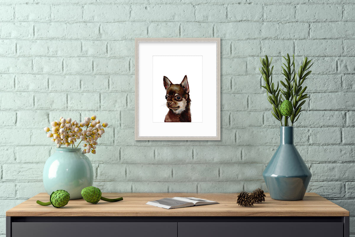An illustration of a Chihuahua dog framed in a silver frame hangs on a mint green brick wall above a wooden console table with an open book and contemporary table decor. 