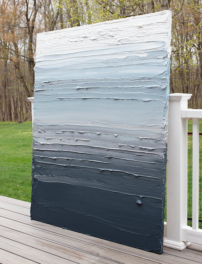 A dark grey blue, light blue, blue green, and white thickly textured abstract painting leaning on a deck outside in natural light by Teodora Guererra.