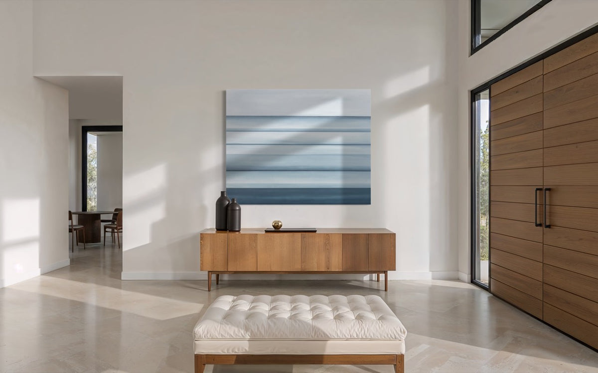 A blue abstract coastal painting hangs on a white wall above a wooden console table in the entryway of a minimalist modern home. 