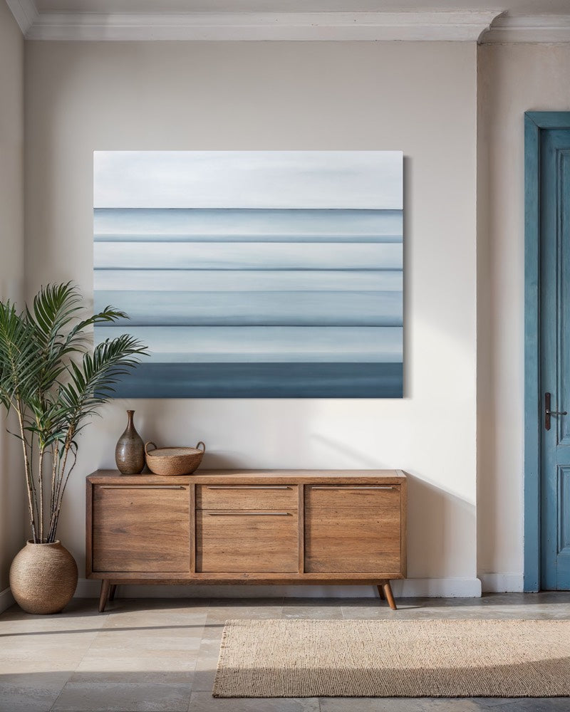 An abstract blue coastal painting hangs above a weathered wooden console table between a floor plant and a blue door. 