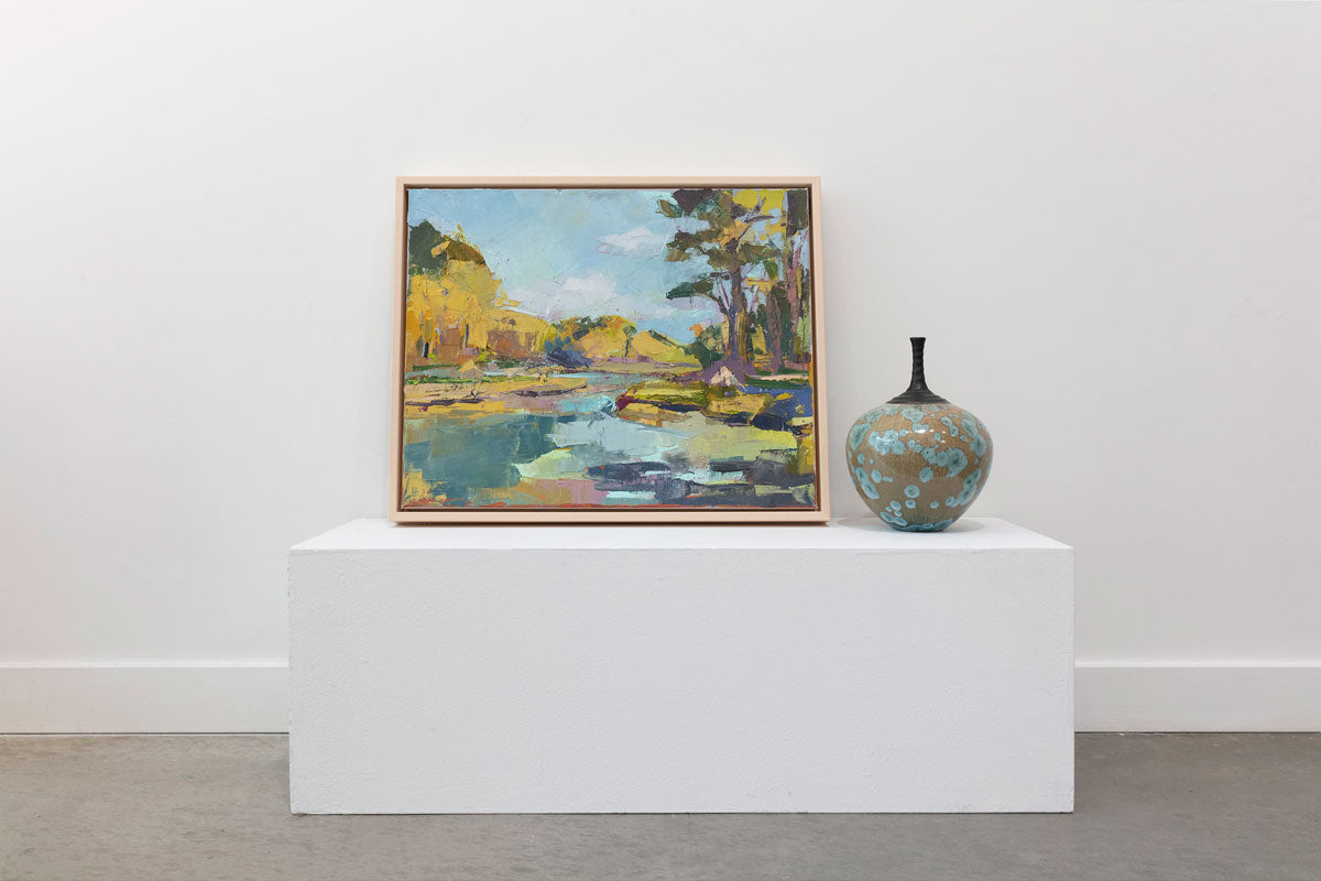 A blue and yellow abstract landscape painting framed in a natural floater frame sits leaning on a white pedestal with a green and ocher ceramic pot.