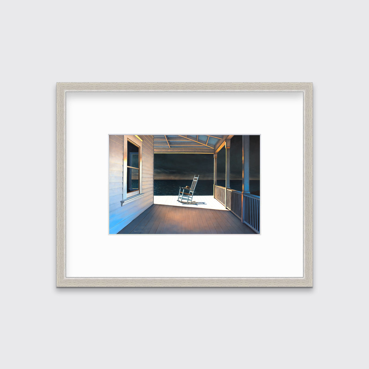 A print of a white rocking chair on a deck of a house with a high contrast light on the chair matted in a silver frame hangs on a white wall.