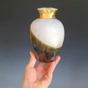 A hand holds a brown and white ceramic vase with a wide gold neck in front of a grey background.