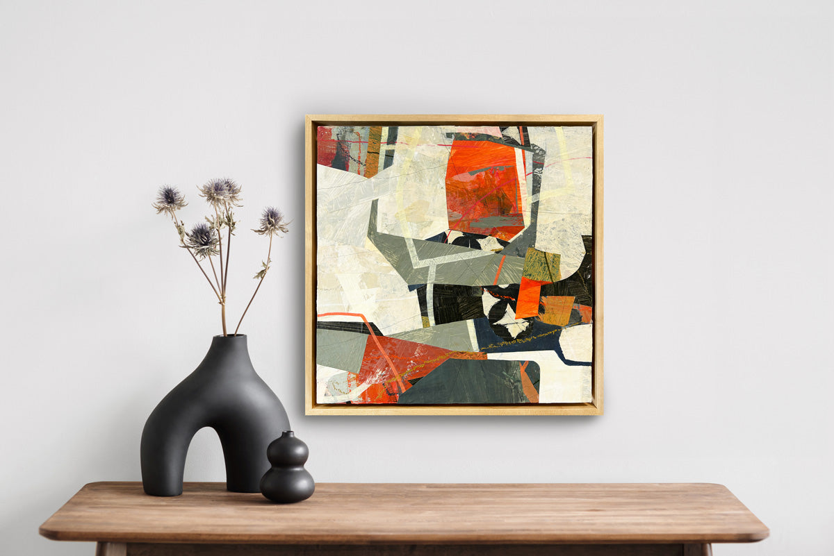 A framed abstract collage artwork featuring geometric shapes in shades of red, beige, and green, with a white background hanging on a white wall.