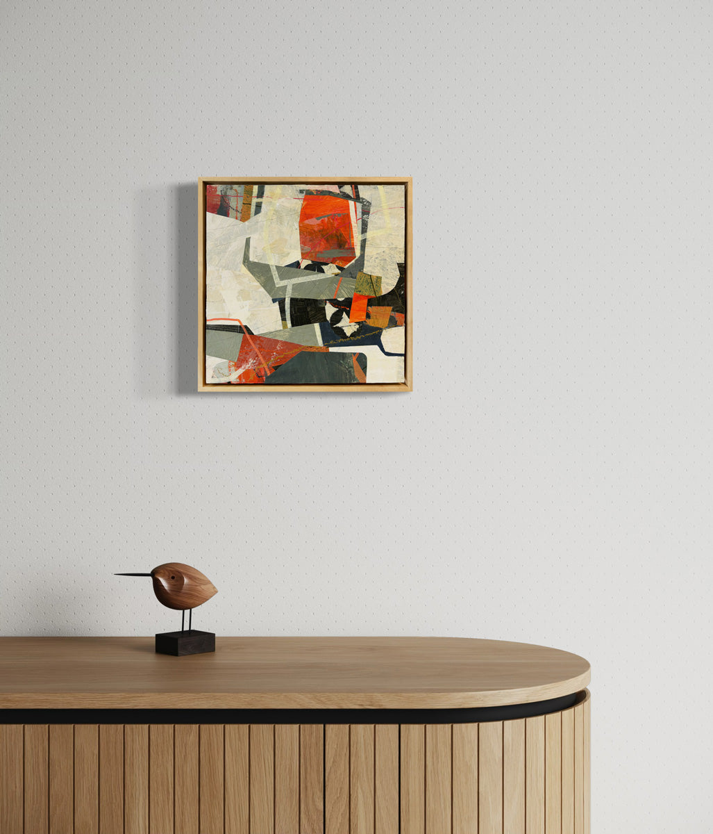 A framed abstract collage artwork featuring geometric shapes in shades of red, beige, and green, with a white background hanging on a white wall.