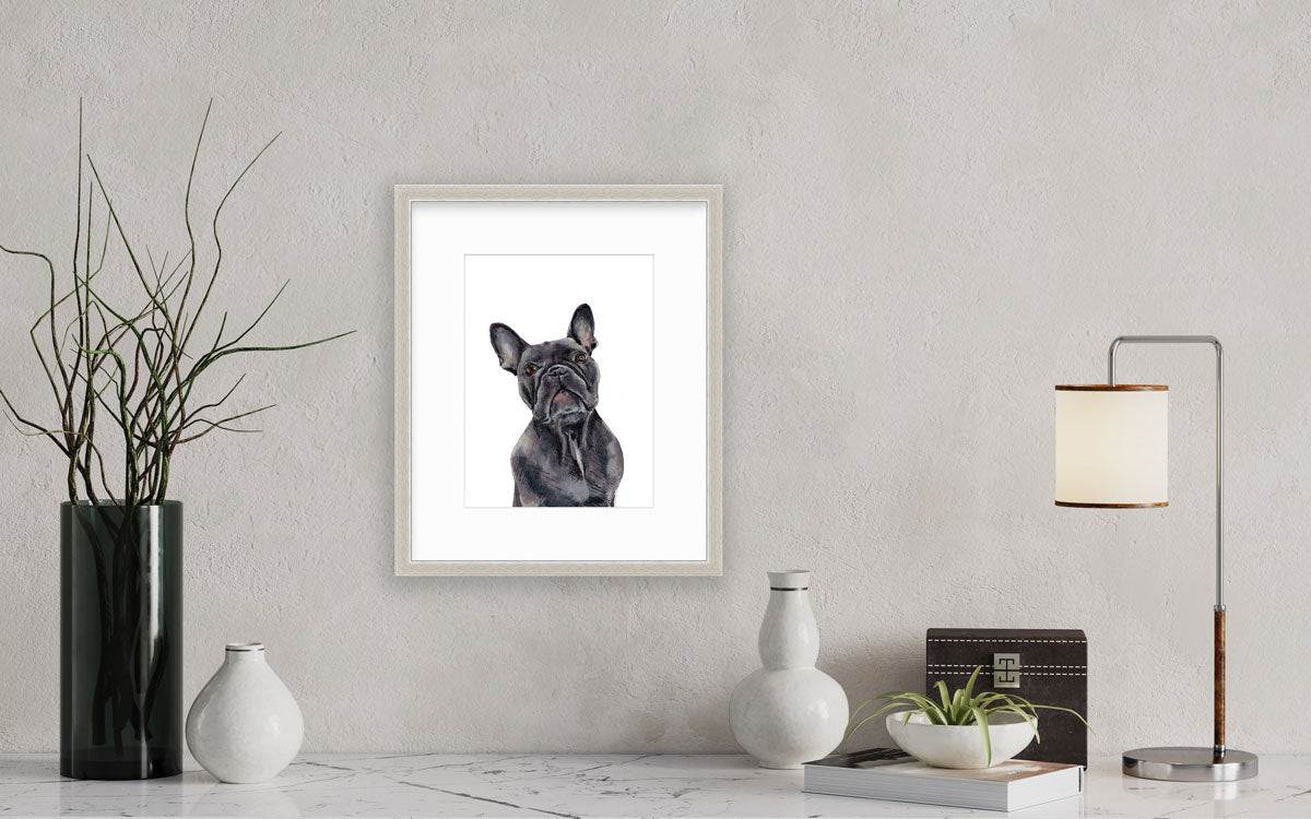 An illustration of a French bulldog framed in a silver frame hangs on a grey textured wall above a marble console table with modern decor and a table lamp. 