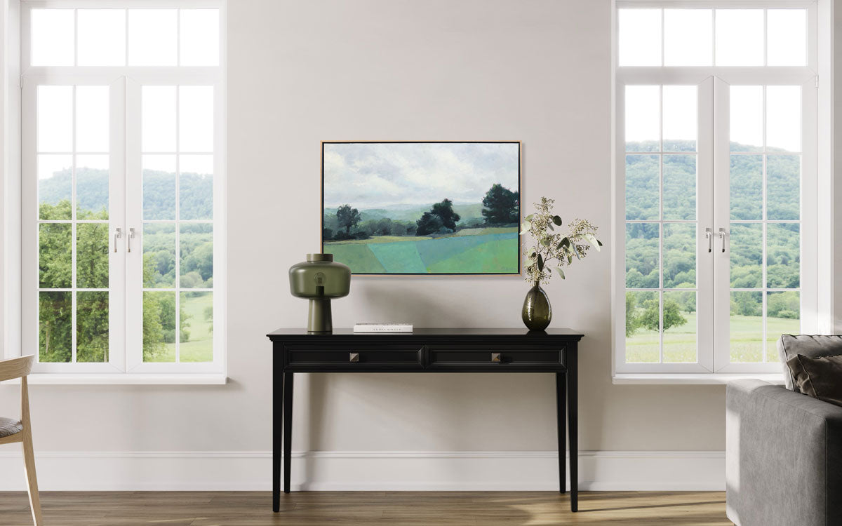A green abstracted landscape painting framed in a wood frame hangs on a white wall between two windows, above a black console table with green table decor. 