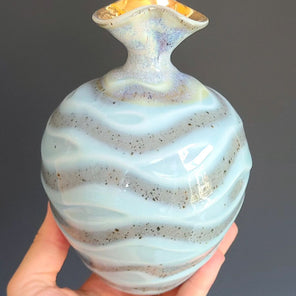 A man's hand holds a glazed ceramic vase with a flared neck finished with gold in front of a grey backdrop. 