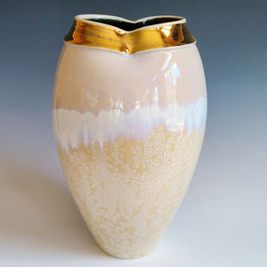 A cream, white, and gold ceramic vessel rests in front of a blue backdrop. 