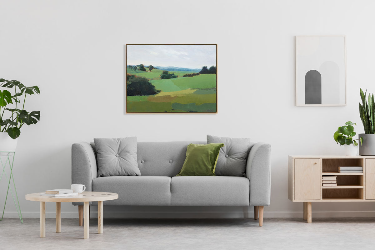 A green abstracted landscape painting hangs on a white wall above a light grey sofa with green accent pillow.