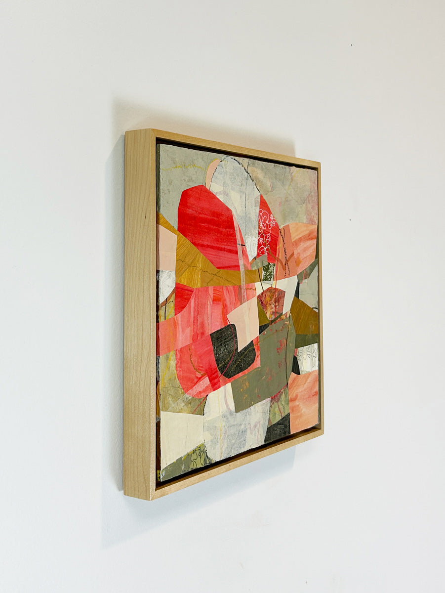 A framed abstract collage artwork featuring geometric shapes in shades of red, beige, and green, with a white background hanging on a white wall, side view.