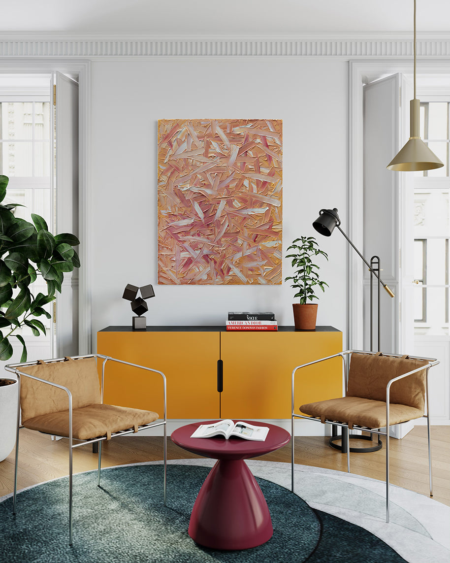 A orange, pink, coral and white thickly textured abstract painting hangs on a white wall above a mid-century yellow orange credenza with a black top. On the credenza is a sculpture, books and potted plant. In front of the credenza are 2 modern chairs with yellow ochre cushions.  A low red table is in front of the chairs and sits on a grey rug. 