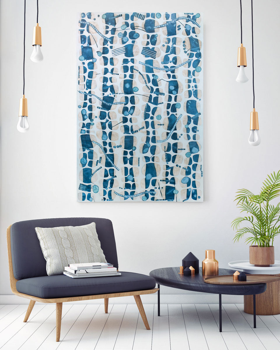 An abstract blue and white painting hangs on a white wall between hanging light fixtures and above a low grey armchair and dark wood coffee table with table decor and a potted plant. 