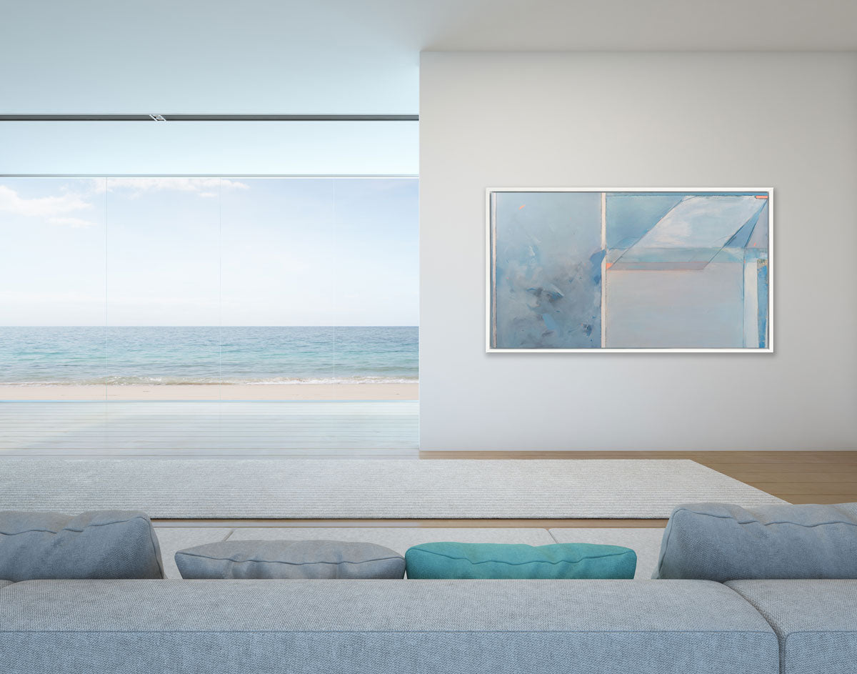 A blue, grey, and pink abstract art print by Kelly Rossetti framed in a white floater frame hangs on a white wall next to a floor-to-ceiling window with view of the beach with a blue and green sofa in the foreground. 