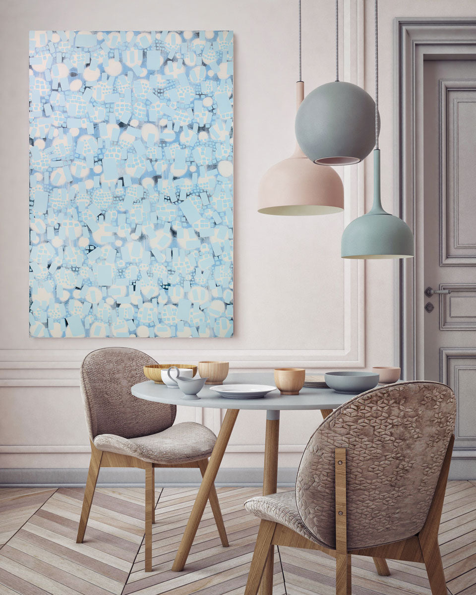 A blue and white abstract painting hangs on a white wall above a modern table and chair and colorful modern hanging lights. 