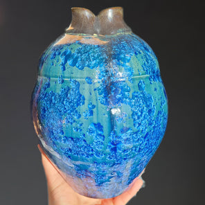 A hand holds a large blue and turquoise glazed ceramic vessel with a brass-toned neck from it's base in front of a grey backdrop. 