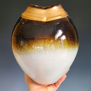 A hand holds a white, yellow, and umber-toned ceramic vase with a wide gold neck in front of a grey background. 