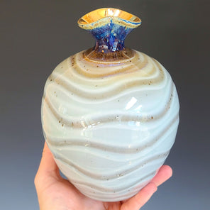 A hand holds a light green and sandy beige ceramic vessel with a flared blue and gold neck in front of a grey gradient background. 